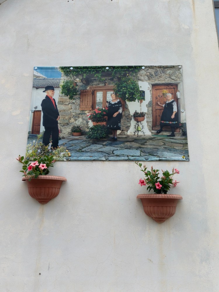 Un'immagine di un pannello affisso su una parete, raffigurante due persone in abiti tradizionali davanti a una casa rustica, con fiori in vasi appesi.