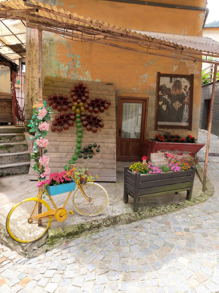 Una bicicletta gialla decorata con fiori colorati accanto a una parete di legno con un disegno floreale e piante in vasi, in un angolo pittoresco di un villaggio.