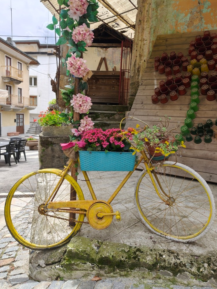Una bicicletta gialla decorata con un cesto blu pieno di fiori colorati, situata in un ambiente pittoresco.