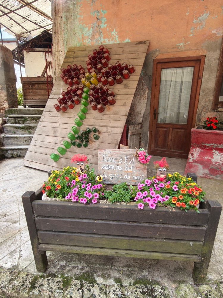 Un mural decorativo che rappresenta un fiore stilizzato realizzato con vasi colorati appesi su una parete, con una fioriera in legno piena di fiori colorati e un cartello con una scritta.