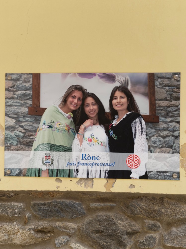 Tre donne in abiti tradizionali, sorridenti, davanti a una parete di pietra. Un cartello con il testo 'Rònc, pais francoprovensal' è visibile in basso.