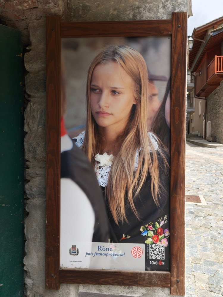 Manifesto che ritrae una ragazza con i capelli lunghi e biondi, vestita con un abito tradizionale, in un ambiente urbano.