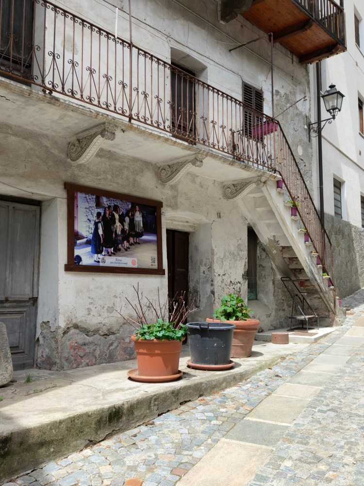 Vista di una strada acciottolata con una vecchia abitazione, un poster appeso al muro e vasi di fiori sul marciapiede.