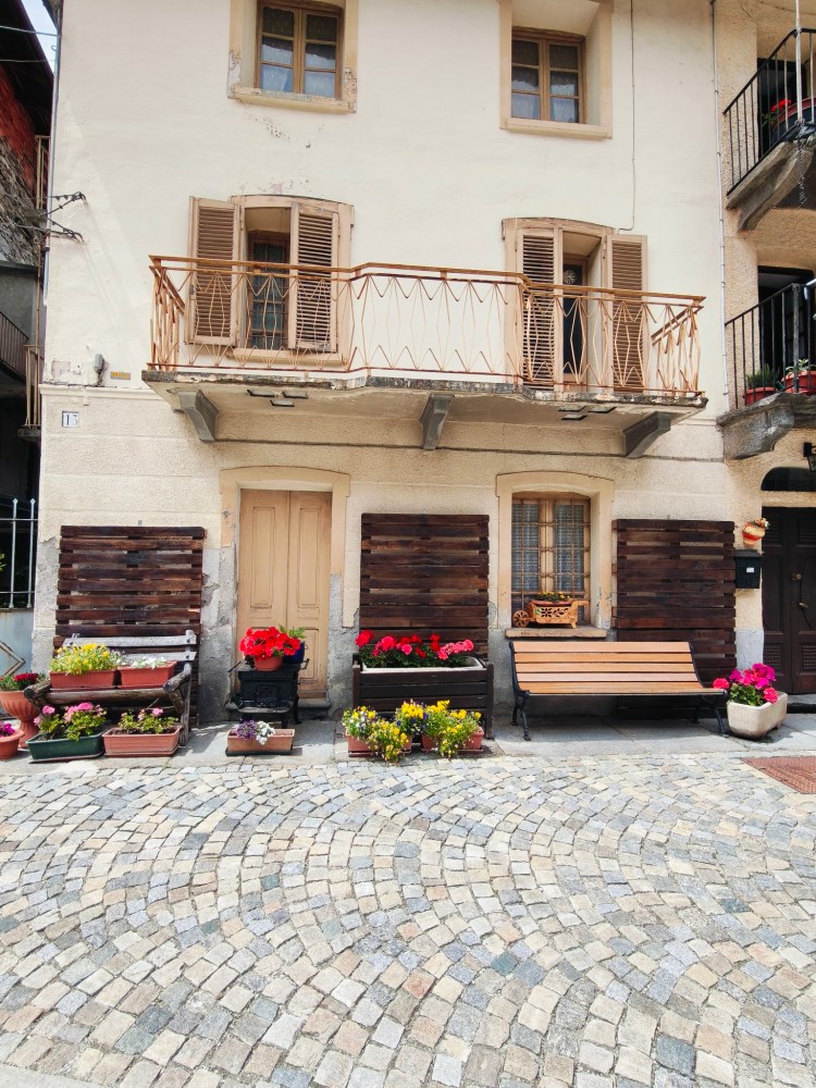 Facciata di un edificio con un balcone e finestre. Fiori colorati in vasi e fioriere lungo la strada acciottolata.