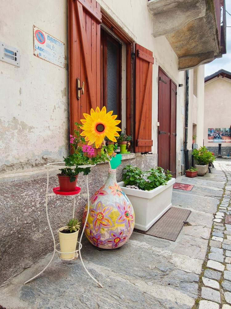 Una fioriera decorata con un girasole e piante colorate davanti a una casa con porte di legno, situata lungo un vicolo acciottolato.