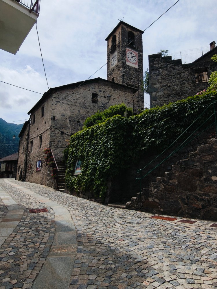 Una strada acciottolata che conduce a un edificio in pietra con un campanile e orologio, circondato da vegetazione verde.