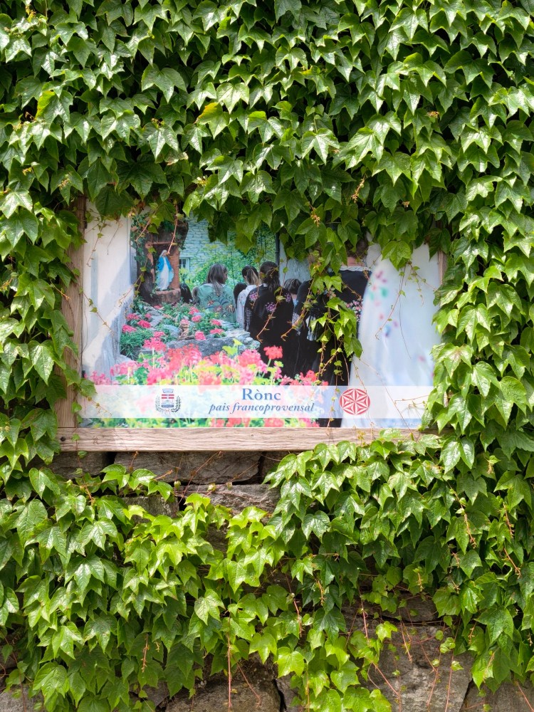 Un pannello affisso su un muro coperto di edera, mostrando un'immagine di persone in un giardino fiorito con il testo 'Rònc paìs francoprovensal'.