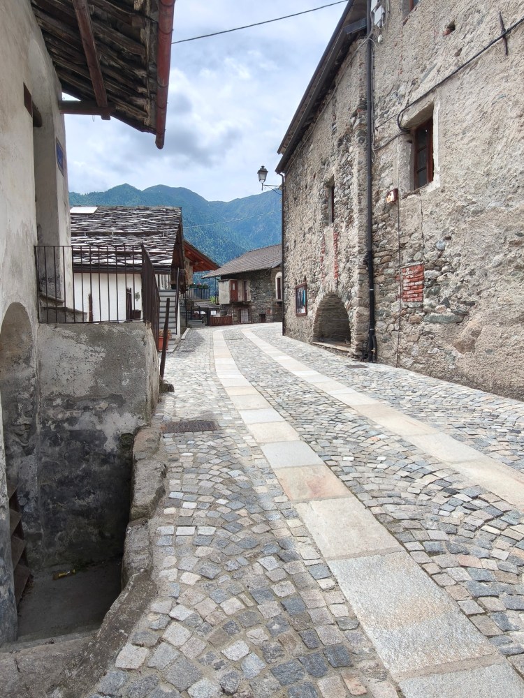 Una strada ciottolata in un villaggio montano, con edifici in pietra e tetti in legno. Sullo sfondo si vedono le montagne e il cielo nuvoloso.
