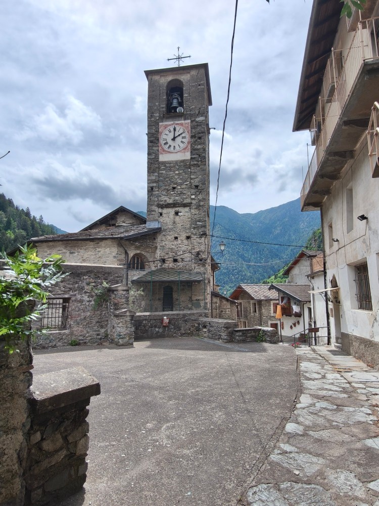 Piazza con un antico campanile in pietra e una torre dell'orologio, circondata da case tradizionali e montagne sullo sfondo.