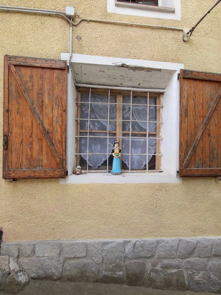 Finestra con sbarre in legno, decorata con una bambola e una statuetta, circondata da persiane di legno su una parete gialla.
