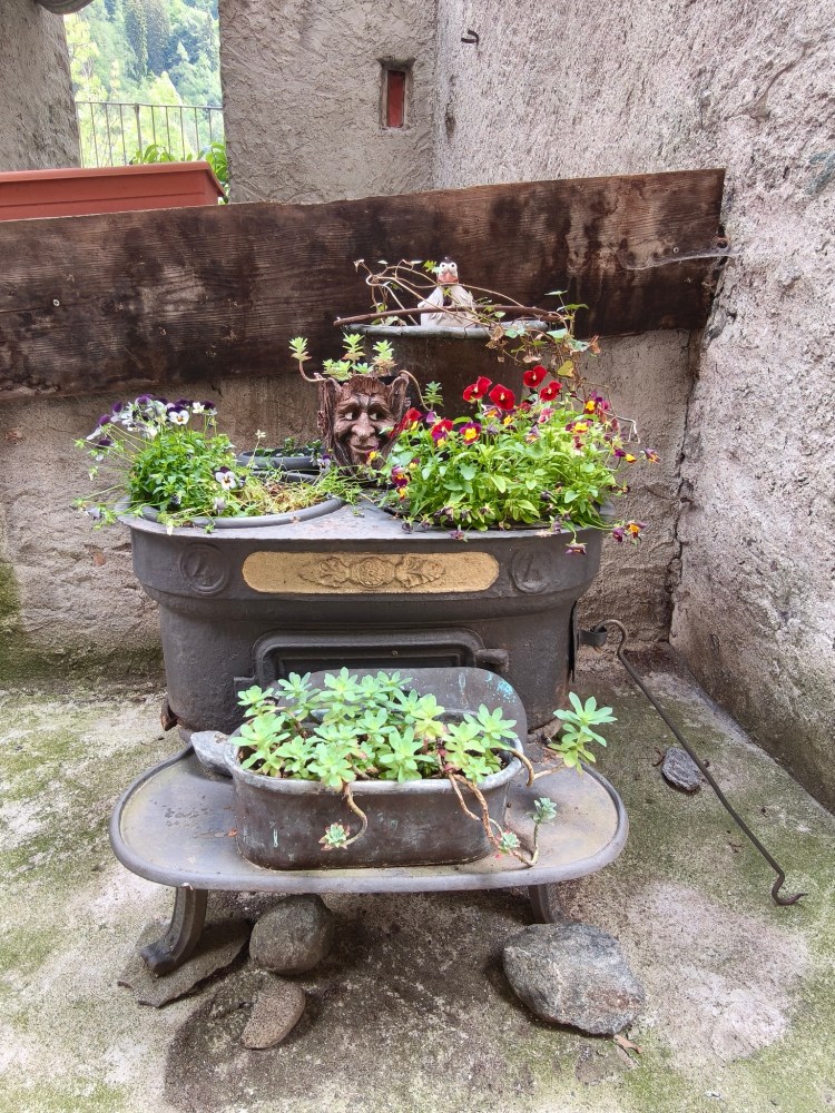 Vasa da fiori decorata con piante variopinte e un volto intagliato, posizionata su una stufa metallica in un ambiente esterno.