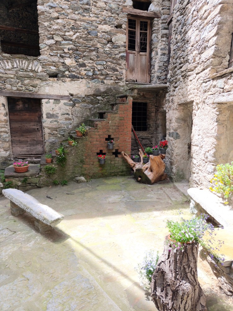 Un cortile all'aperto con pareti in pietra, scale in mattoni, piante in vaso e un tronco di legno su un lato.
