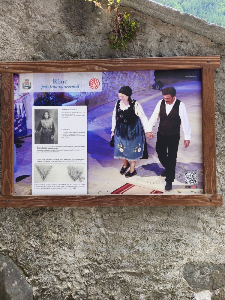 Un pannello informativo di legno con immagini e testo descrittivo sulla tradizione del paese francoprovenzal di Rònc, presentando persone in abiti tradizionali che partecipano a una danza.