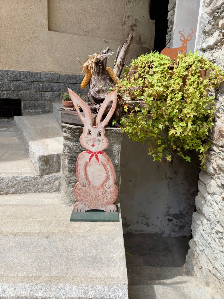 Un coniglio di legno decorativo con un fazzoletto rosso, posizionato su una scalinata accanto a una pianta verde e a del legno.