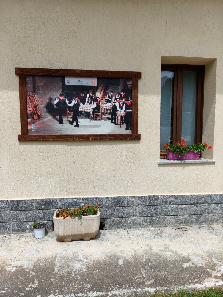 Un'insegna fotografica appesa a una parete che ritrae un gruppo di persone in abiti tradizionali che partecipano a un evento culturale, con piante fiorite in vaso sotto.