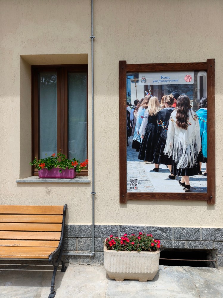 Affisso su una parete esterna, un poster che mostra un gruppo di persone in abiti tradizionali, mentre una finestra con fiori colorati è visibile accanto.
