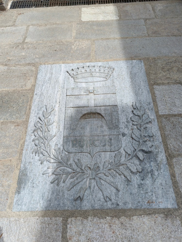 Scultura in pietra grigia con il simbolo del comune di Runcus, con un castello e una corona, circondata da un ramoscello.