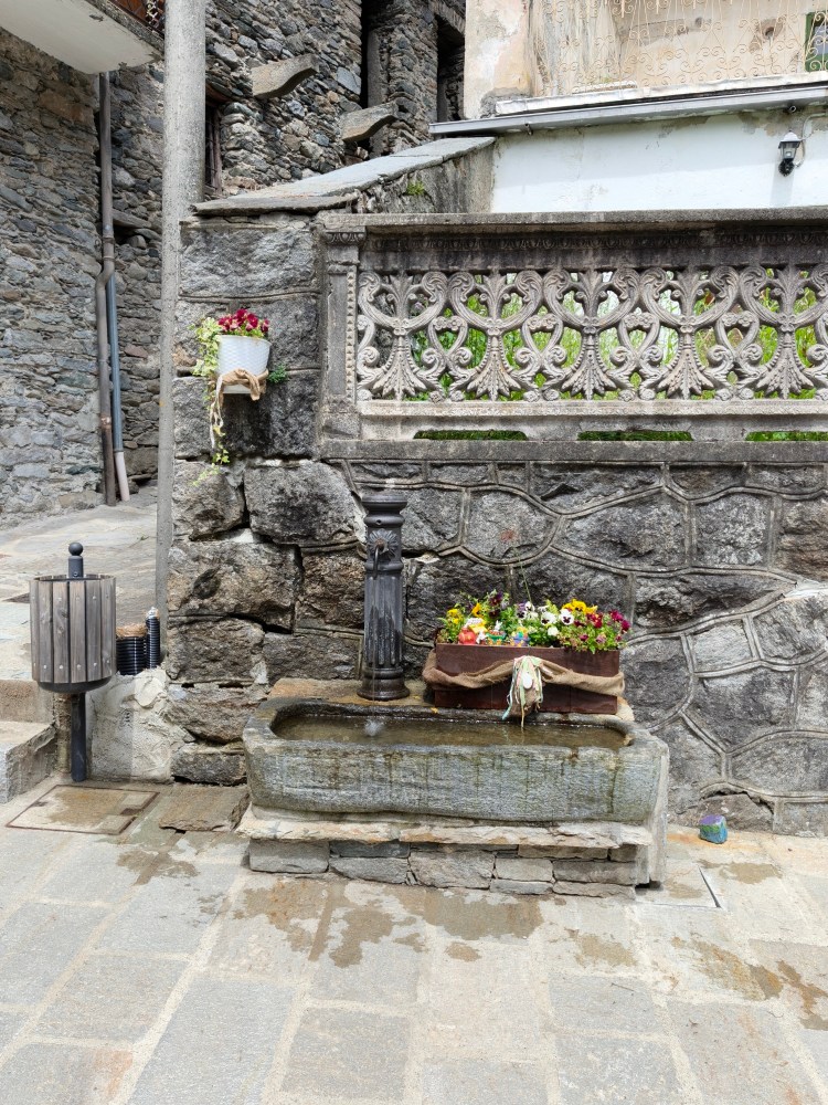 Una fontana in un villaggio con un lavabo in pietra e fiori colorati in un vaso accanto.