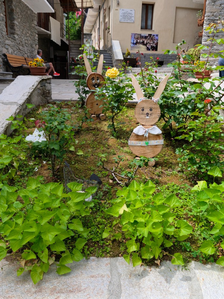Giardino con decorazioni a forma di coniglio in legno tra piante verdi e fiori, con una persona seduta su una panchina sullo sfondo.