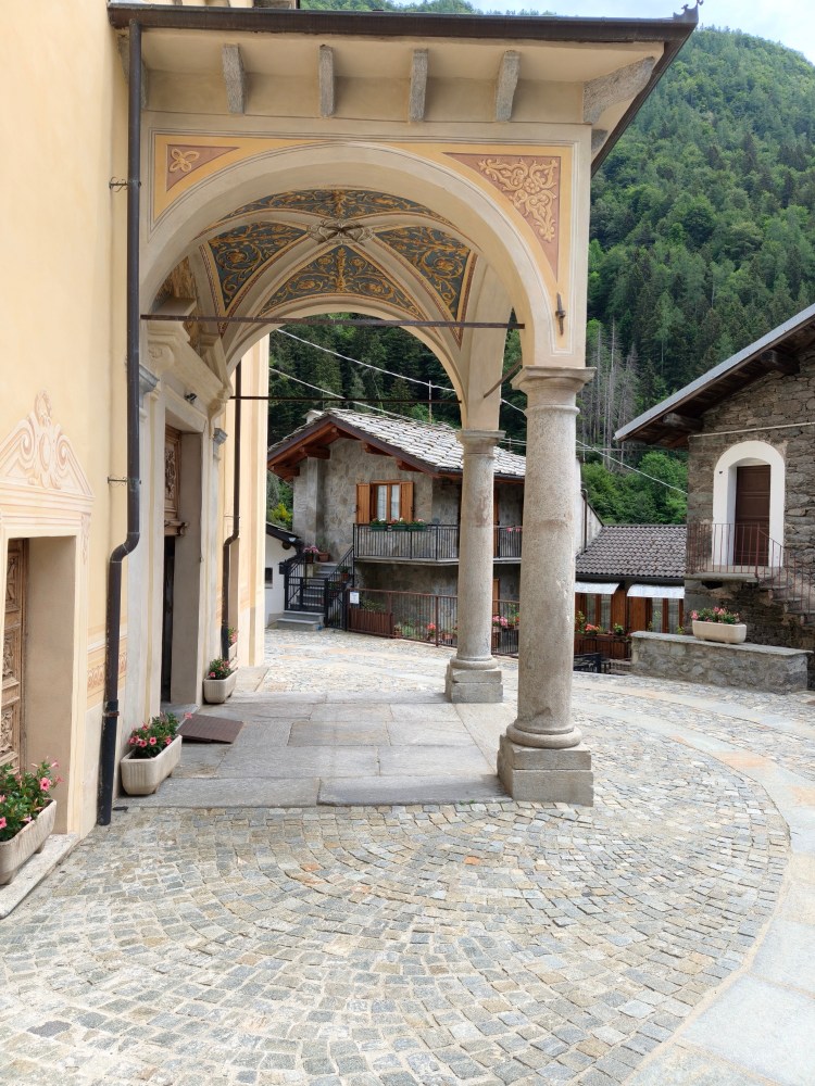 Arco architettonico decorato che conduce a una strada acciottolata in un villaggio di montagna, circondato da edifici tradizionali e vegetazione.