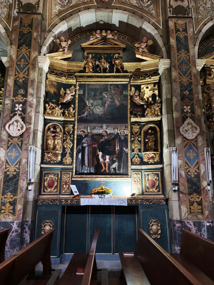 Interno di una chiesa con un altare decorato, affreschi e statue, panchine in legno visibili.