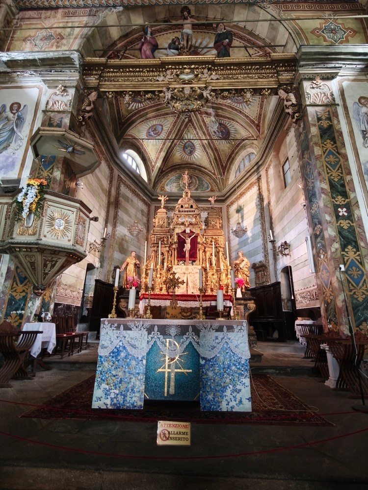 Interno di una chiesa con un altare ornato e dettagli decorativi. Alle pareti ci sono affreschi e decorazioni artistiche. Sullo sfondo si vede un soffitto affrescato e figure religiose dorate.