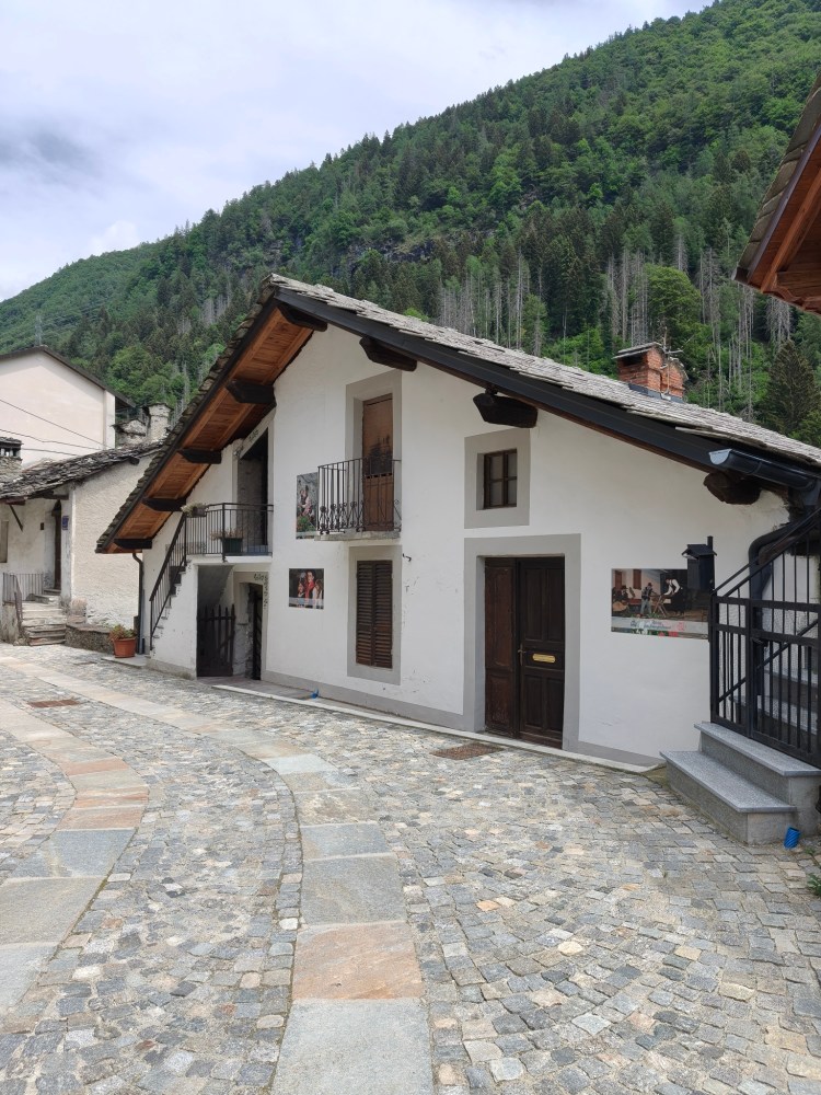 Una casa in stile tradizionale situata in un villaggio montano, con un tetto spiovente e balconi in legno, circondata da una strada acciottolata e colline verdeggianti in lontananza.