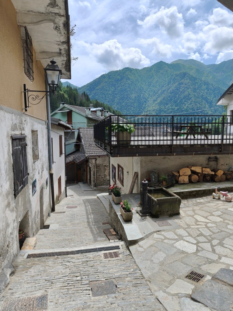Una strada acciottolata in un villaggio montano, con case storiche e una fontana in pietra. Sullo sfondo, montagne verdi e nuvole nel cielo.