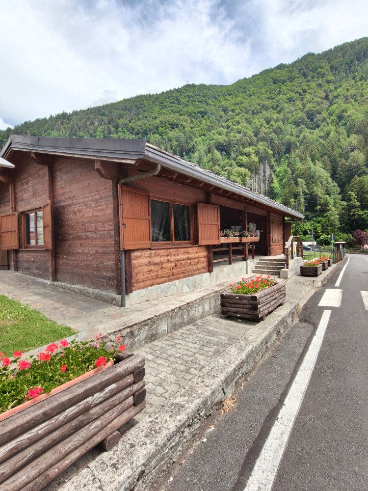 Casa in legno con finestre e persiane, circondata da un giardino con fiori e una strada asfaltata, sullo sfondo una collina verde.