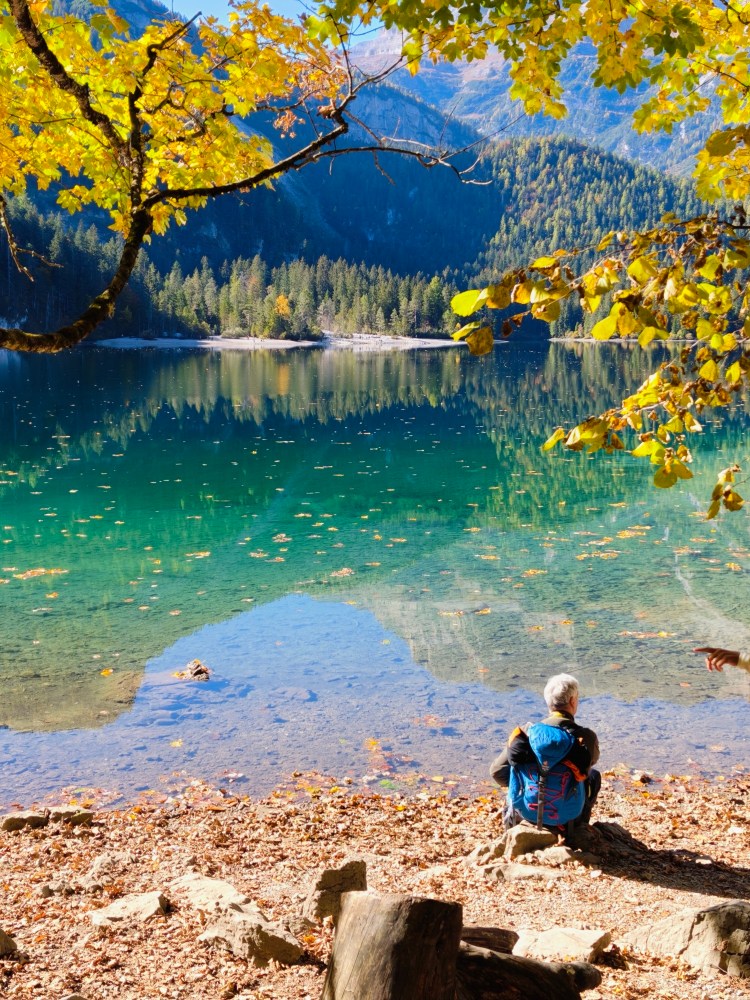Una persona seduta sulla riva di un lago cristallino circondato da alberi con foglie autunnali colorate.