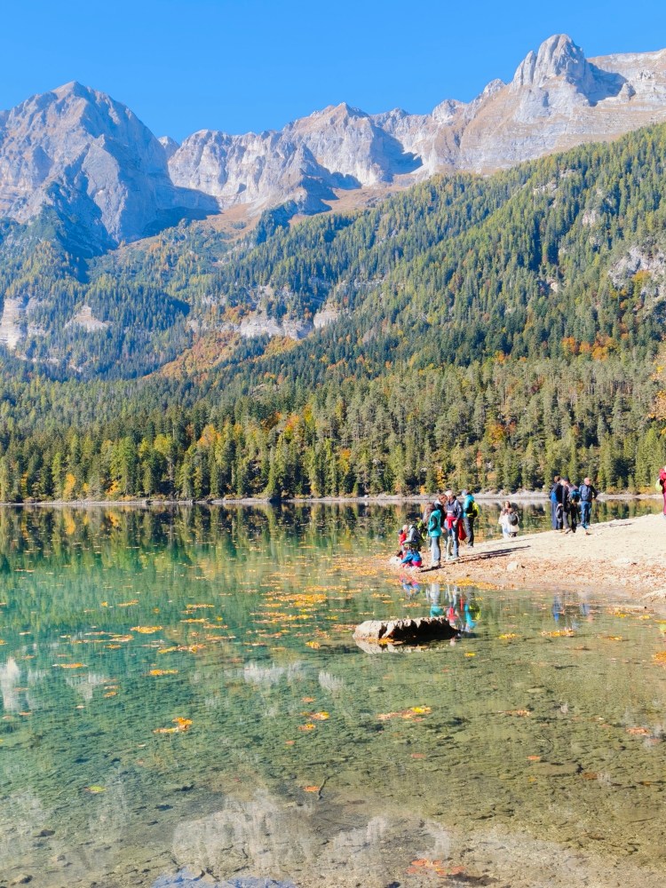 Un gruppo di persone vicino a un lago cristallino circondato da montagne e alberi verdi, con foglie autunnali galleggianti sulla superficie dell'acqua.