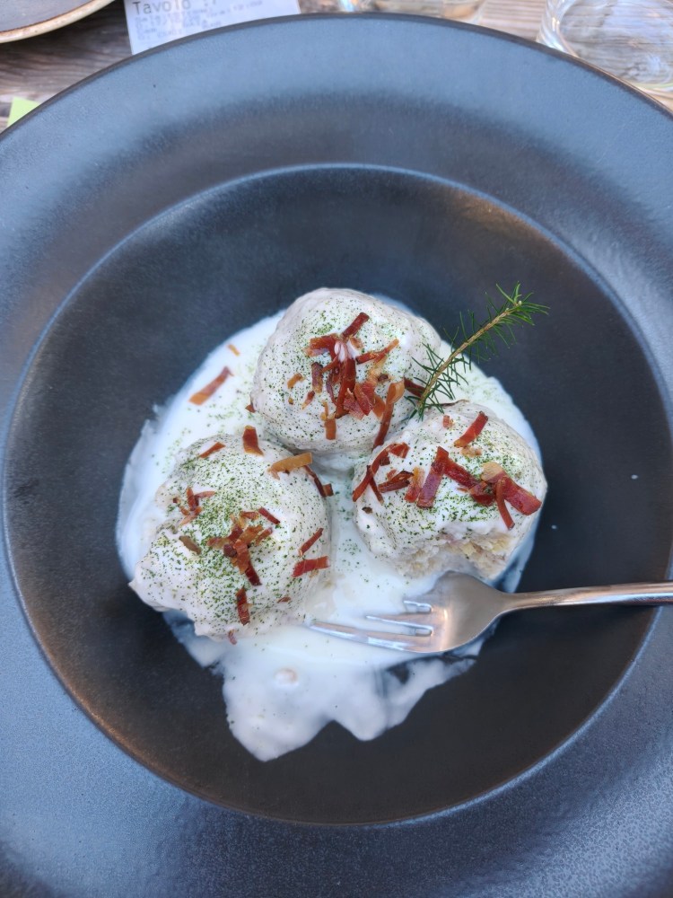 Piatto di tre palline di dessert guarnite con pezzetti di pancetta, erba aromatica e salsa bianca, servito in un piatto nero.