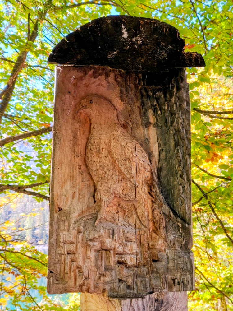 Scultura di legno di un uccello, incisa su un tronco, con sfondo di foglie verdi e blu del cielo.