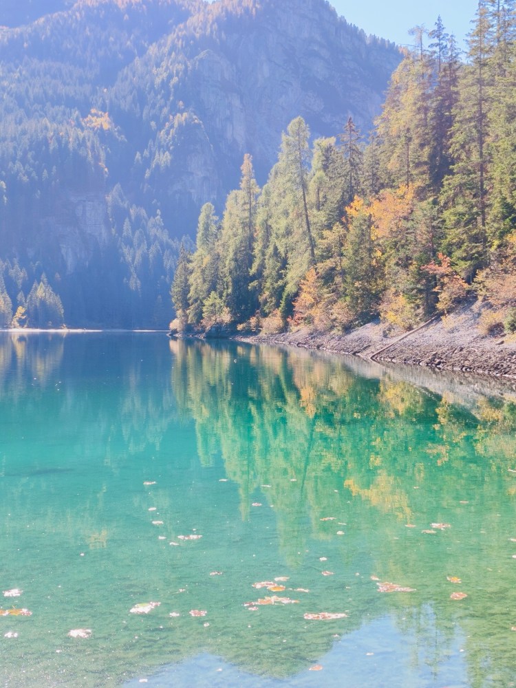Un lago cristallino circondato da alberi verdi e autunnali, con riflessi delle montagne sullo sfondo.