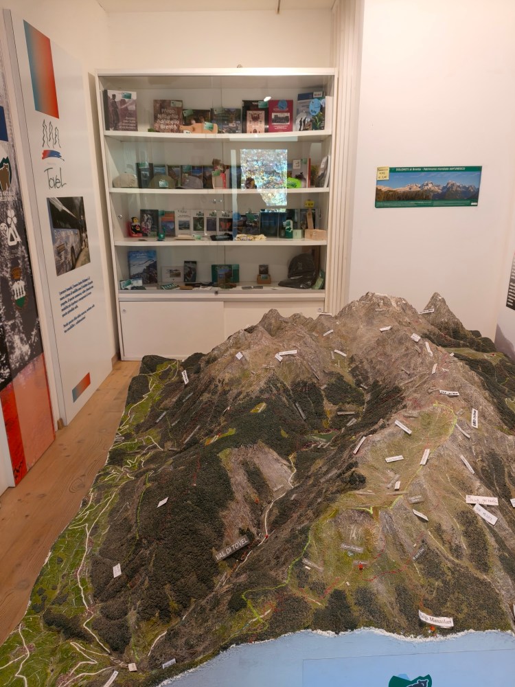 Modello in miniatura delle montagne con etichette informative, all'interno di un museo. Sullo sfondo, una vetrina con oggetti e brochure relative al territorio montano.