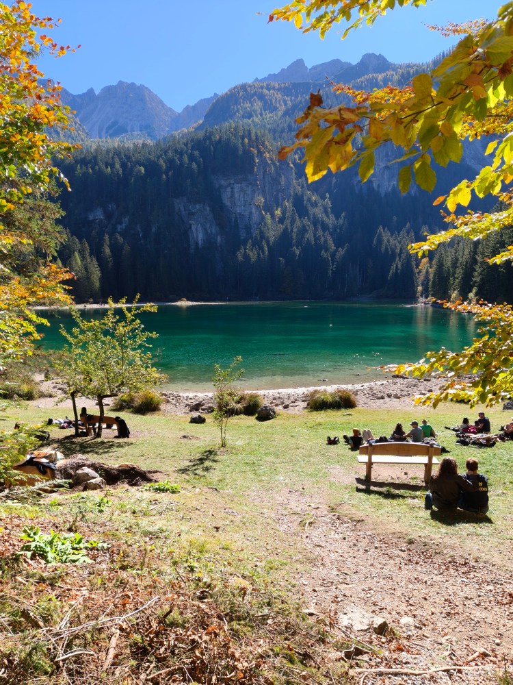 Un lago con acqua verde smeraldo circondato da alberi e montagne, con persone che si rilassano sulla riva e seduti su panchine.