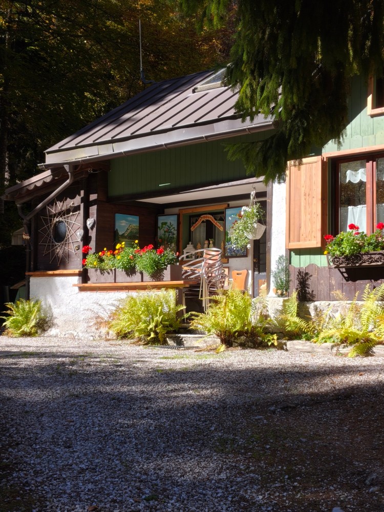 Facciata di una casa di montagna con fiori colorati sul portico e sedie impilate, circondata da alberi e piante verdi.