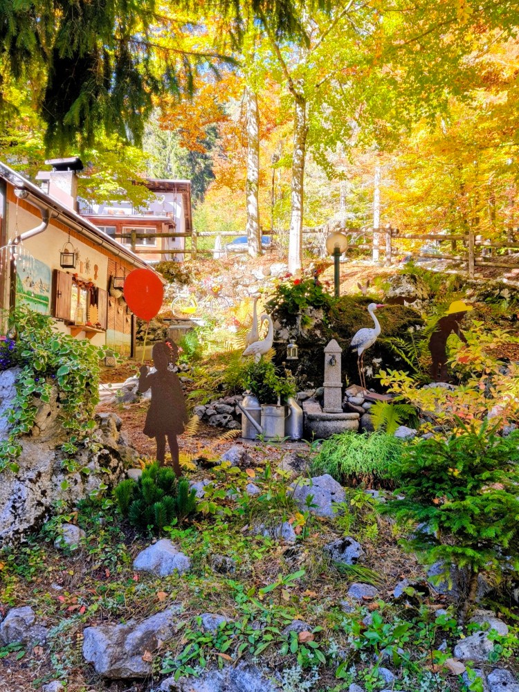 Giardino autunnale con alberi colorati, una casa rustica e decorazioni, inclusa una figura di una ragazza con un palloncino rosso e sculture di fenicotteri.