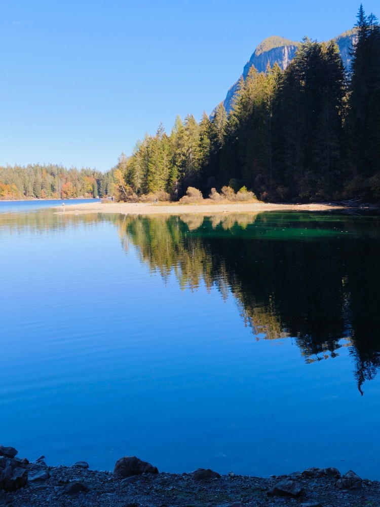 Panorama di un lago tranquillo circondato da foreste verdi e montagne sotto un cielo azzurro.