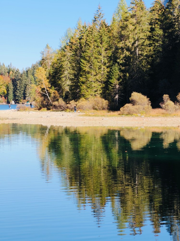 Un paesaggio sereno con un lago che riflette alberi verdi e una riva sabbiosa. Alcune persone passeggiano lungo il lago sotto un cielo blu.