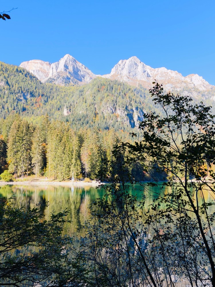 Vista di montagne imponenti sullo sfondo di un lago tranquillo, circondato da alberi verdi e autunnali sotto un cielo blu.