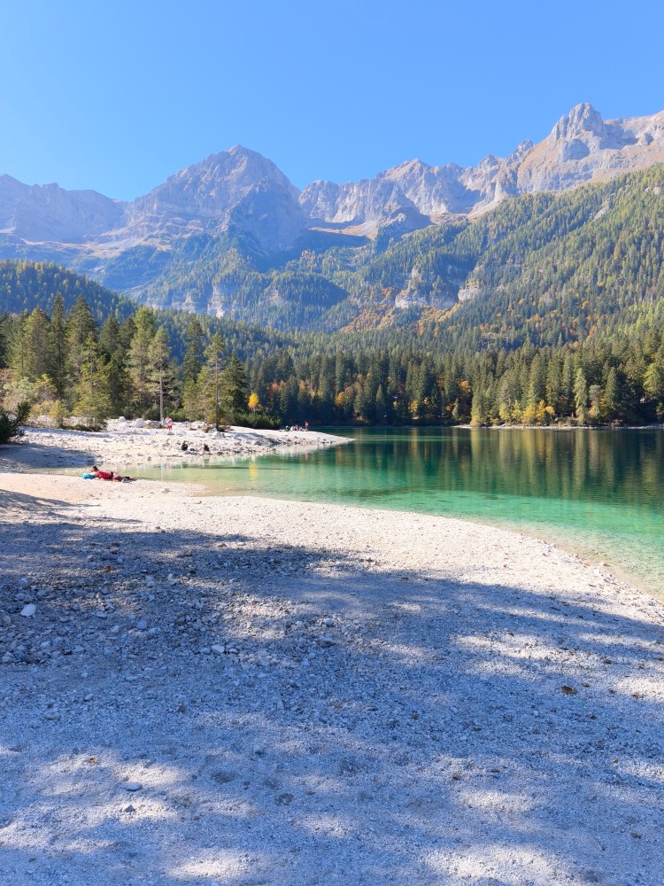 Una vista panoramica di un lago circondato da montagne, con alberi verdi e una spiaggia di ciottoli.
