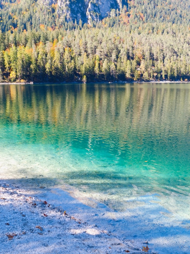 Un lago cristallino circondato da alberi verdi e foglie autunnali, con il riflesso delle montagne sulla superficie dell'acqua.