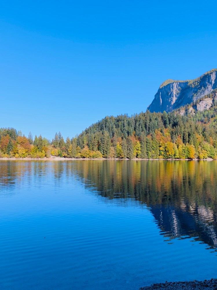 Lago sereno circondato da alberi colorati nel periodo autunnale, con montagne sullo sfondo e cielo blu.