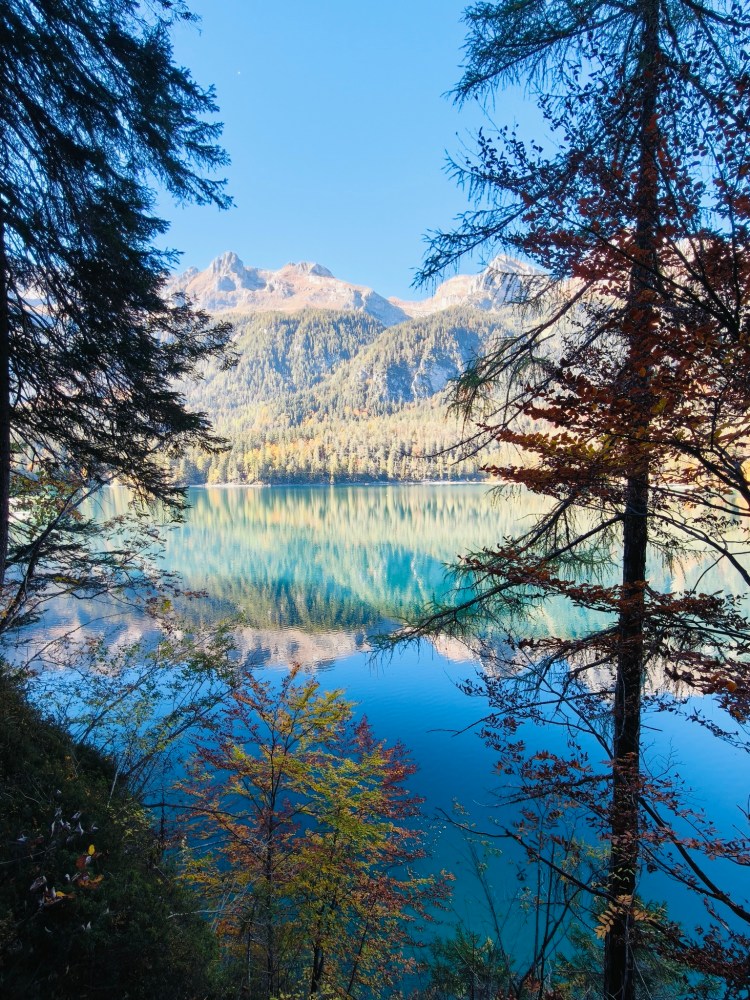 Lago tranquillo circondato da alberi e montagne sullo sfondo sotto un cielo azzurro.