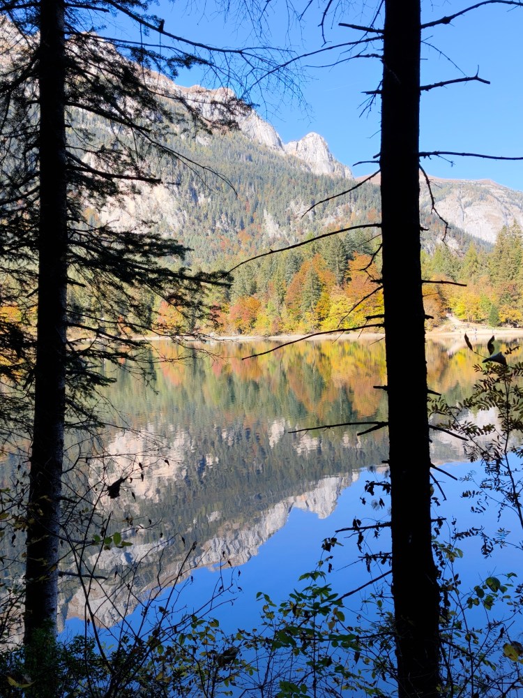 Vista di un lago circondato da alberi, con riflessi delle montagne sullo specchio d'acqua e colori autunnali.