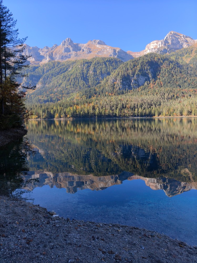 Panorama di un lago tranquillo circondato da montagne e foreste, con riflessi delle montagne nell'acqua.