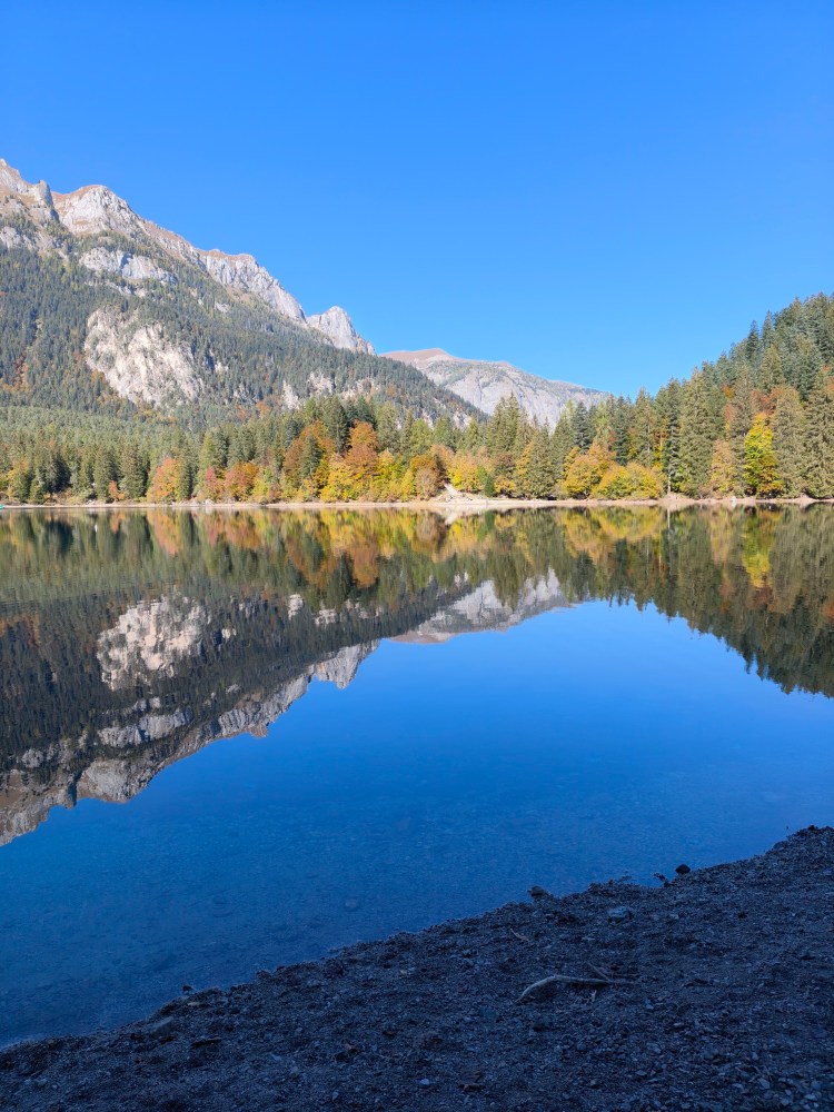 Vista di un lago circondato da montagne e alberi colorati in autunno, con un cielo azzurro e riflessi sull'acqua.