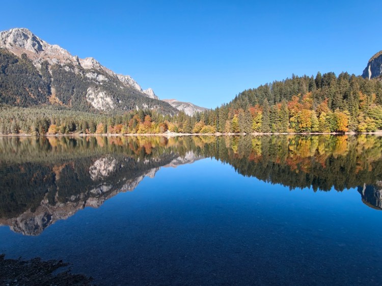 Una vista panoramica di un lago tranquillo circondato da montagne e foreste. Le cime montuose sono riflesse sull'acqua limpida, sotto un cielo sereno e blu. Gli alberi mostrano foglie autunnali variopinte.