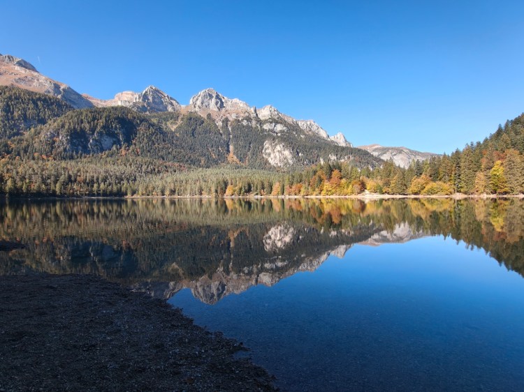 Un lago tranquillo incastonato tra le montagne, riflettendo il cielo blu e gli alberi circostanti. Le cime montuose sono parzialmente coperte di vegetazione autunnale.